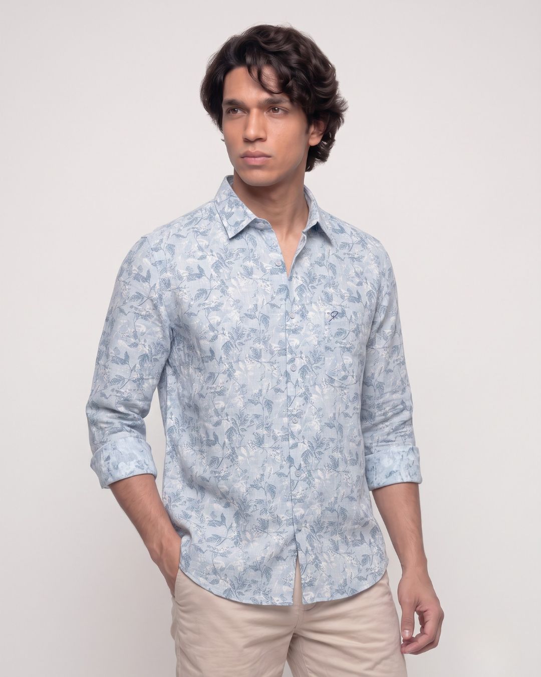 A man wearing a light blue and white patterned long-sleeve button-down shirt with beige trousers, standing against a plain light gray studio background.