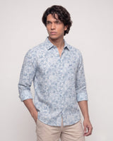 A man wearing a light blue and white patterned long-sleeve button-down shirt with beige trousers, standing against a plain light gray studio background.