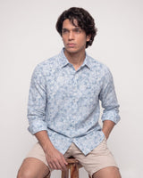 A man wearing a light blue and white floral patterned long-sleeve button-down shirt and beige shorts, sitting on a wooden stool against a plain light gray background.