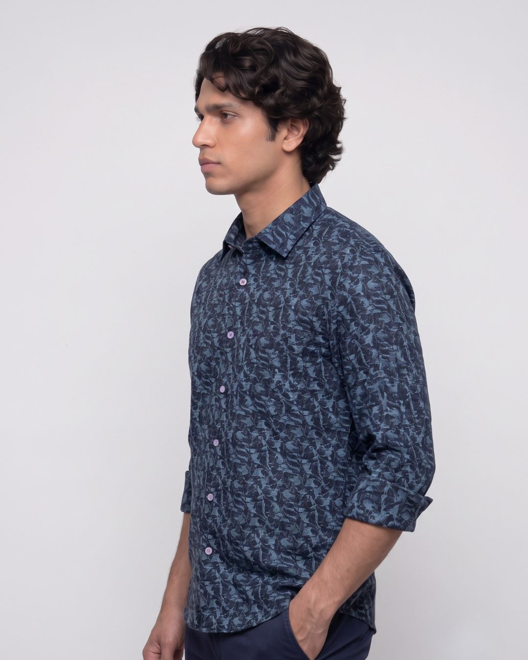 A man wearing a dark blue patterned button-down shirt with rolled-up sleeves, shown in profile against a plain light gray background.