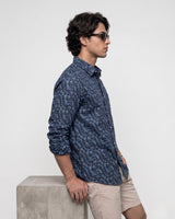A man in profile wearing a navy blue long-sleeve button-up shirt with a light beige abstract pattern, paired with beige shorts, standing next to a concrete block against a light gray background.