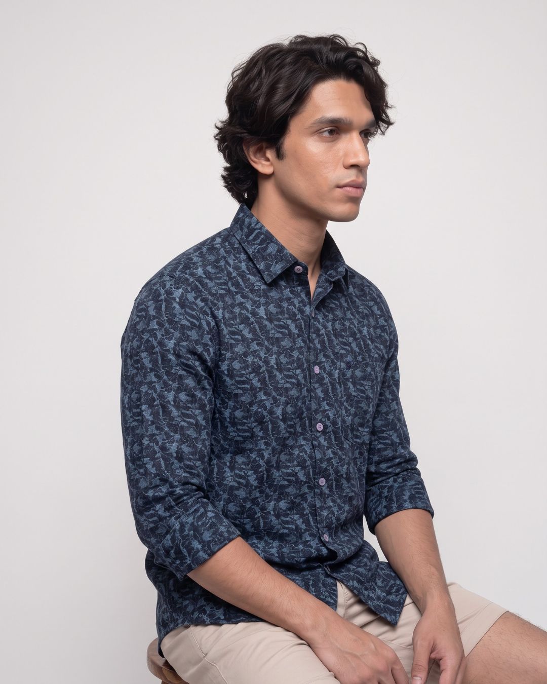 A man wearing a dark navy blue patterned long-sleeve button-down shirt and beige shorts, seated against a plain light gray background.