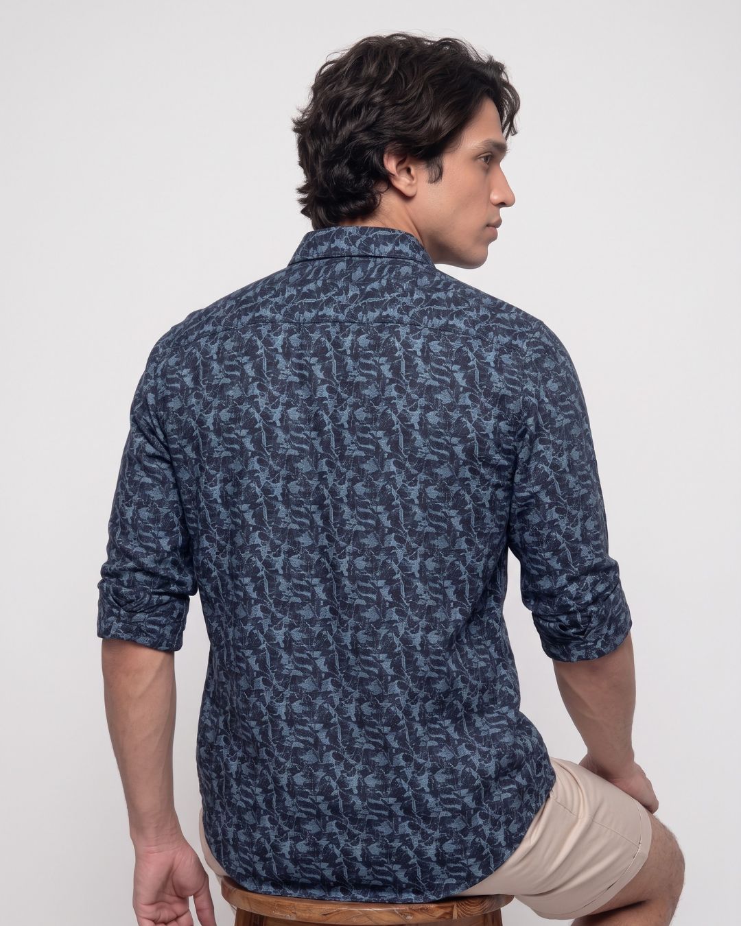 A man wearing a navy blue patterned button-down shirt and beige shorts, shown from the back and side profile against a plain white studio background.