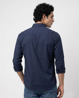 A man viewed from the back wearing a navy blue long-sleeve button-down shirt with rolled-up sleeves and blue jeans, set against a plain light gray background.