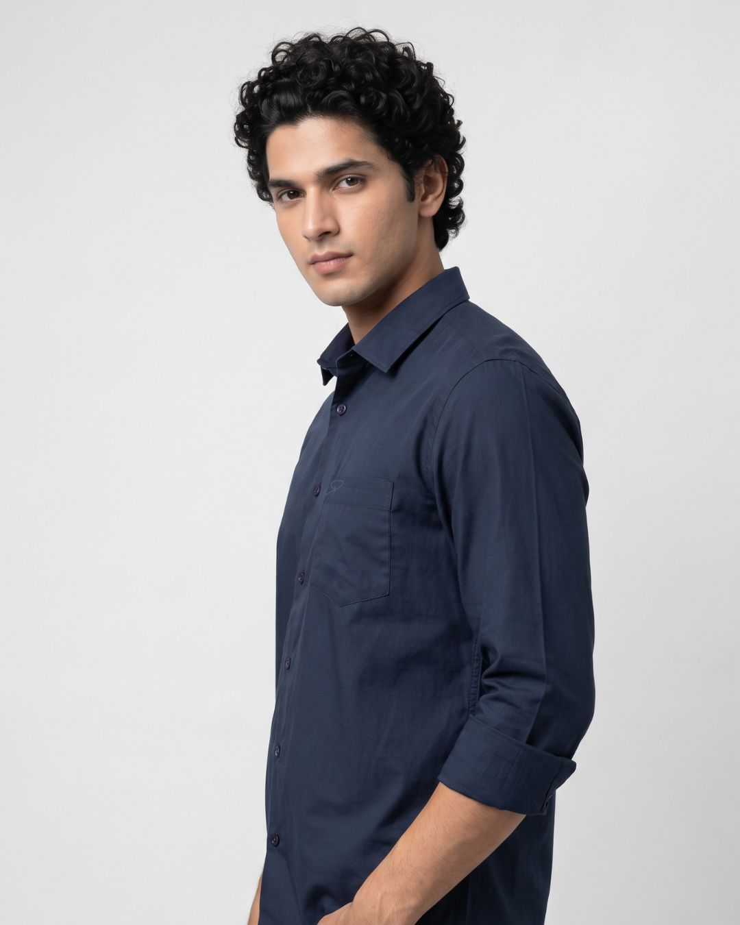 A man wearing a dark navy blue long-sleeve button-down shirt, shown from a side profile against a light gray studio background.