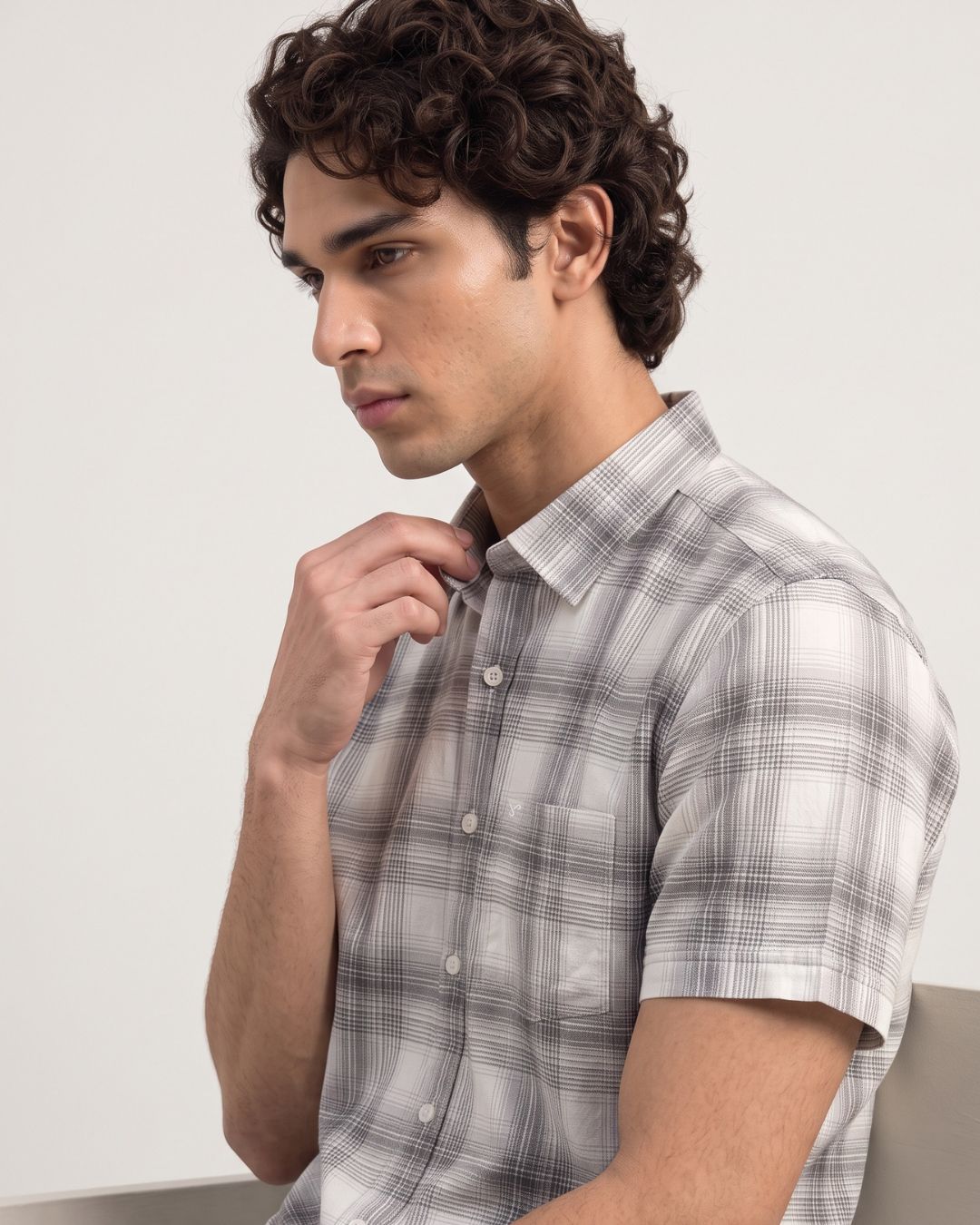 A man wearing a short-sleeve plaid button-up shirt in gray, white, and black, shown in a side profile against a light gray background.