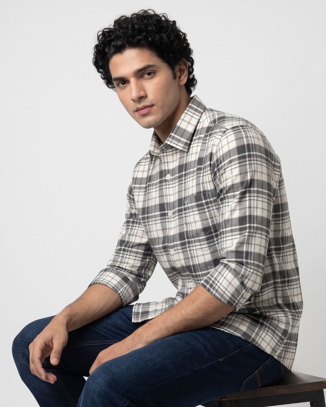 A man wearing a long-sleeve plaid button-up shirt in gray, white, and black, paired with dark blue jeans, sitting against a light gray studio background.