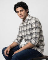 A man wearing a long-sleeve plaid button-up shirt in gray, white, and black, paired with dark blue jeans, sitting against a light gray studio background.