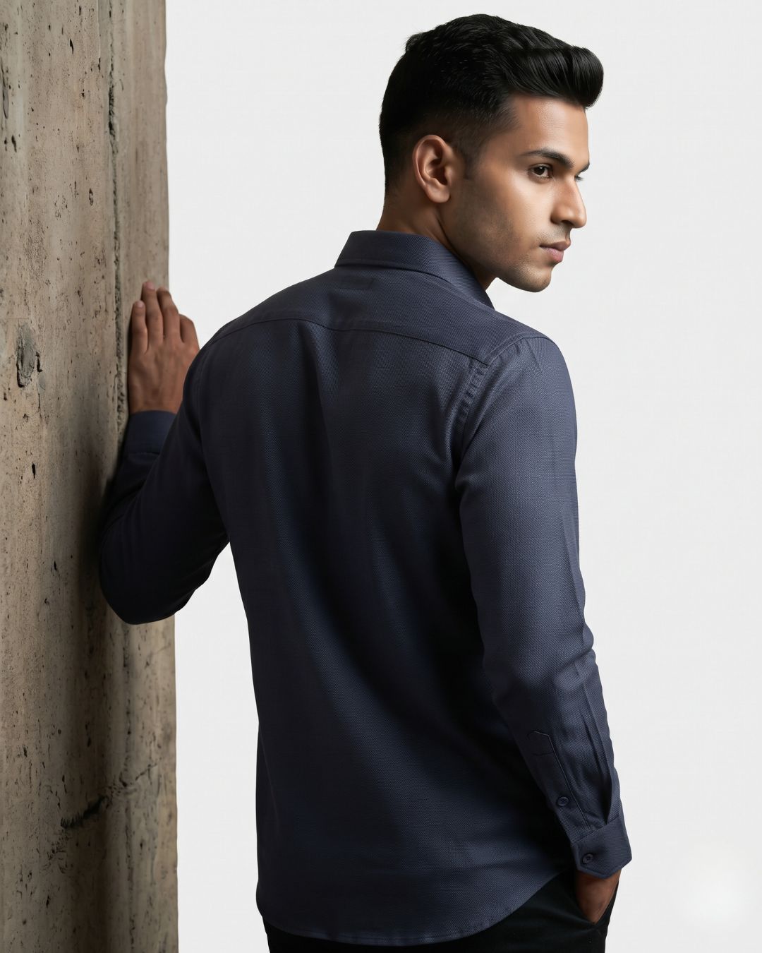 A man wearing a dark navy blue long-sleeve button-up shirt, shown from a three-quarters back view as he leans against a textured concrete pillar.