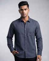 A man wearing a dark slate blue long-sleeve button-down shirt with a single chest pocket, posed against a neutral gray studio background.
