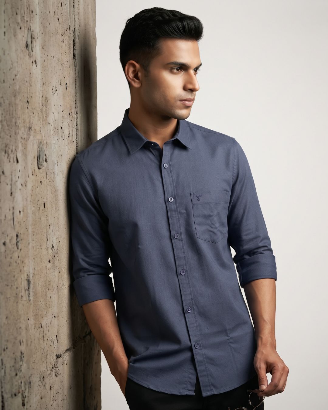 A man wearing a dark slate blue long-sleeve button-down shirt with rolled-up sleeves, leaning against a textured concrete pillar.