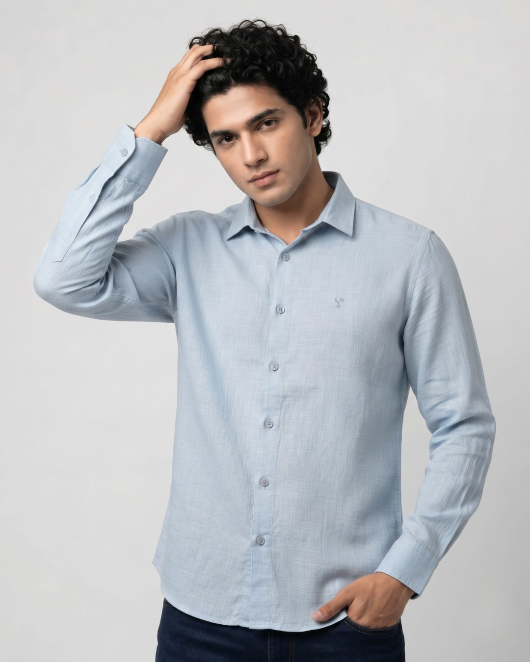 A man wearing a light blue long-sleeve linen button-down shirt and dark blue jeans, posing against a light gray studio background.