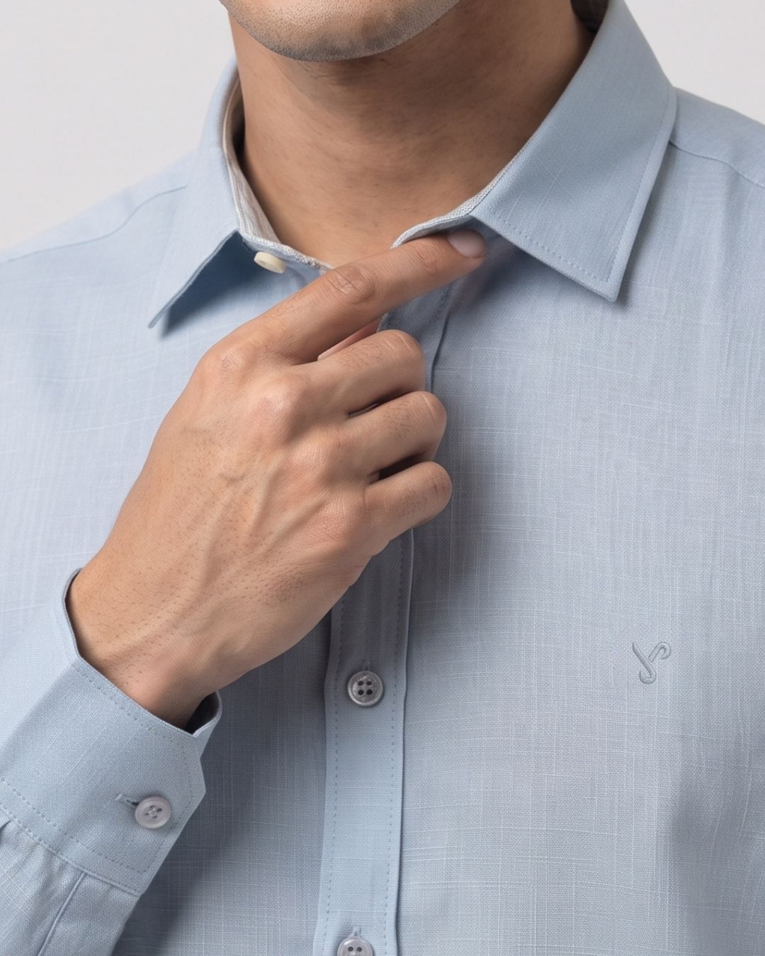 A close-up of a man wearing a light blue textured dress shirt, adjusting the collar with his right hand. The shirt features a subtle grid texture and a small embroidered logo on the left chest.