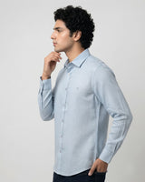 A man wearing a light blue long-sleeve button-down shirt, shown in profile against a plain light gray studio background.