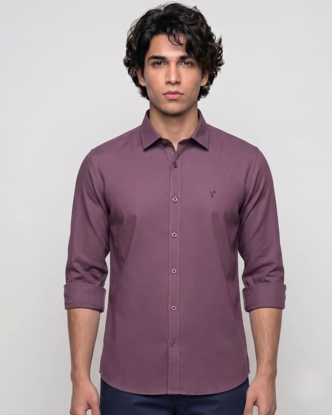 A man wearing a long-sleeve button-down shirt in a muted plum purple color, paired with dark navy pants, standing against a light gray studio background.