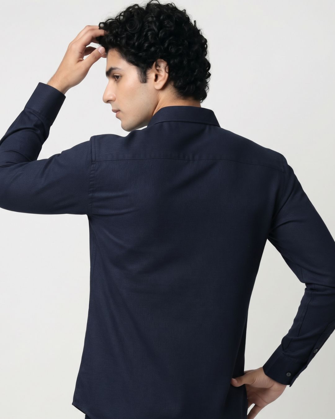 A man wearing a dark navy blue long-sleeve button-up shirt, shown from a three-quarters back view against a plain light gray studio background.