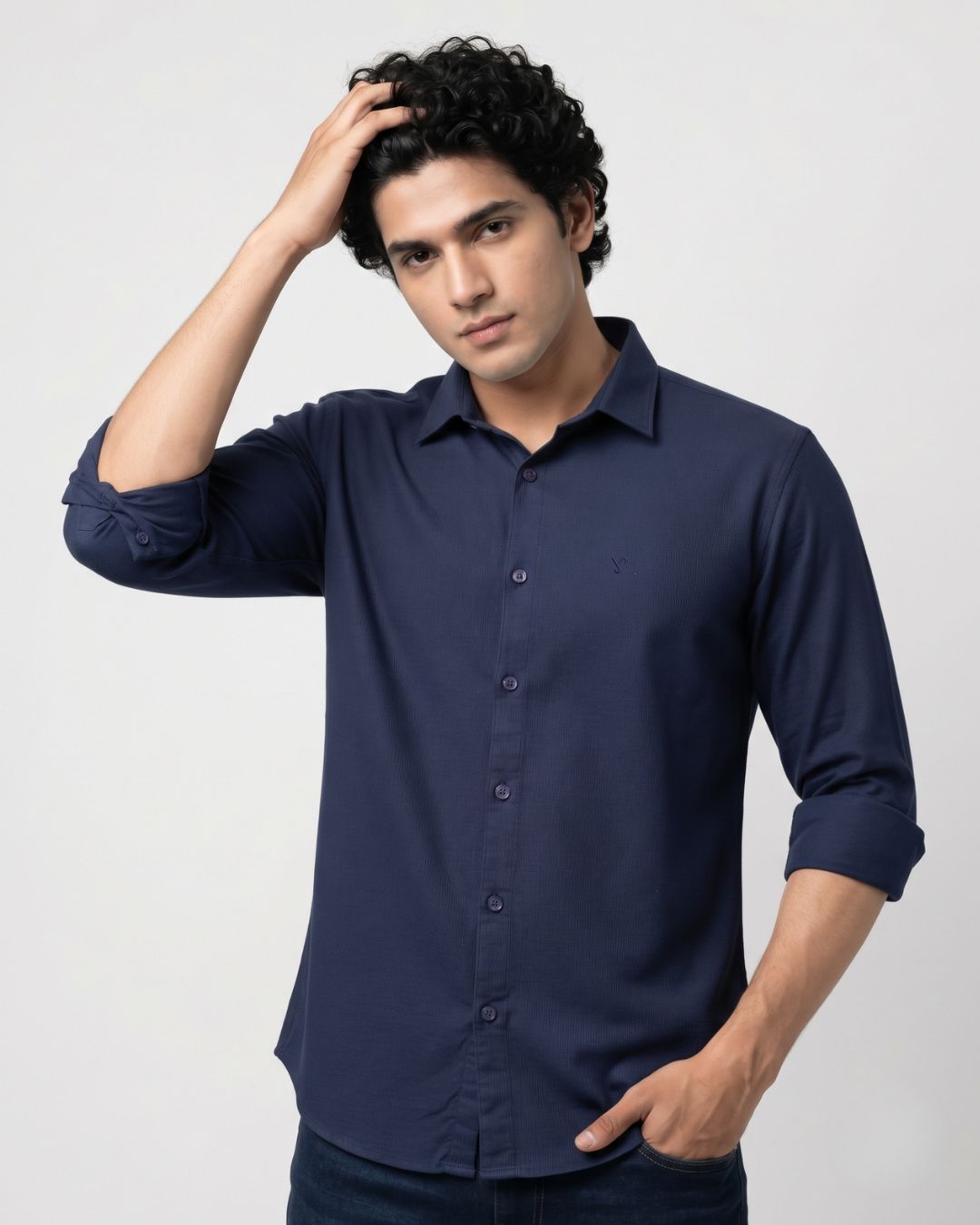 A man wearing a dark navy blue long-sleeve button-down shirt, posing with one hand on his hair against a light gray studio background.