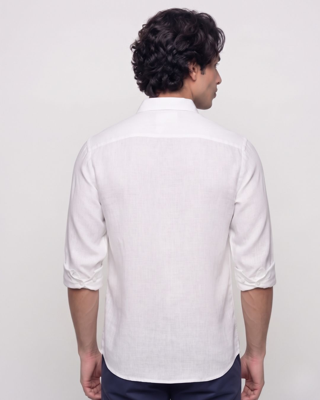A man viewed from the back wearing a white linen shirt with rolled-up sleeves and dark trousers, set against a plain light gray studio background.