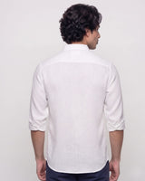 A man viewed from the back wearing a white linen shirt with rolled-up sleeves and dark trousers, set against a plain light gray studio background.