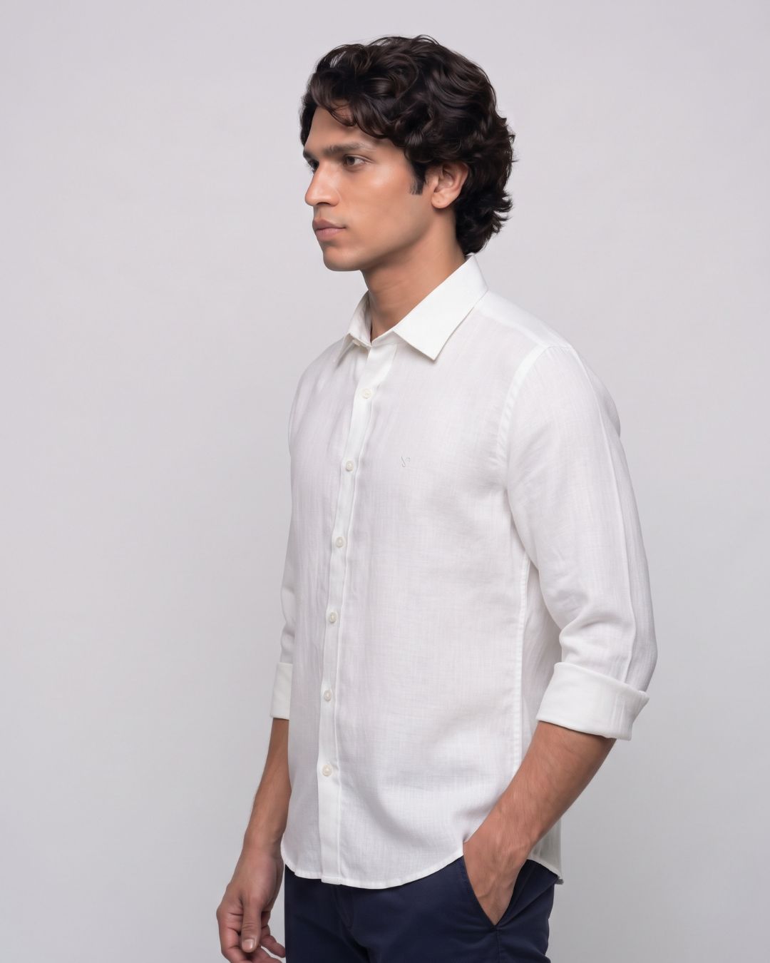 A man wearing a white long-sleeve linen button-down shirt with rolled-up sleeves, paired with dark navy trousers, standing against a light gray studio background.