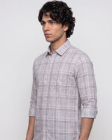 A man wearing a light gray and white plaid button-down shirt with rolled-up sleeves, standing against a plain light gray background.
