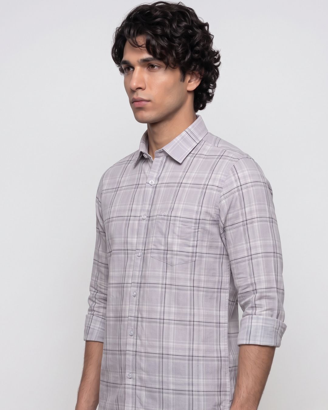 A man wearing a light gray and white plaid button-down shirt with rolled-up sleeves, standing against a plain light gray background.