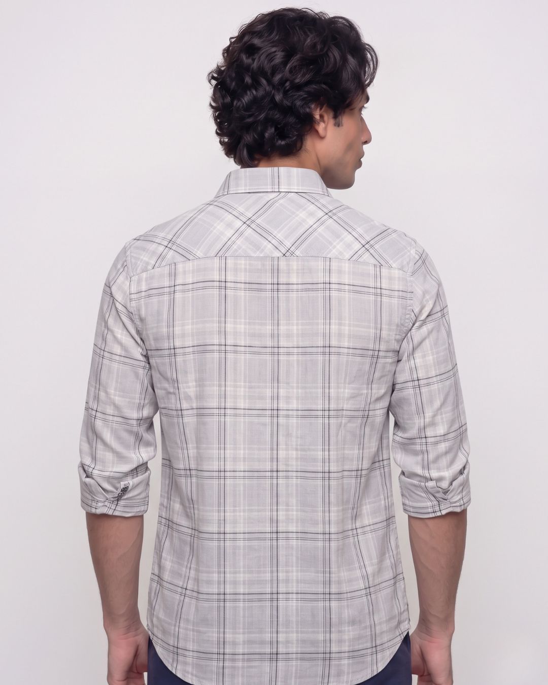 A man viewed from the back wearing a light gray and white plaid button-down shirt with rolled-up sleeves, set against a plain light gray background.