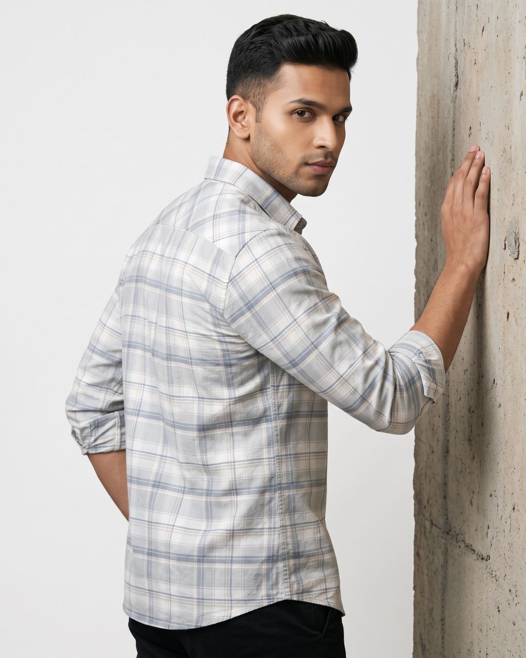 A man wearing a light gray and white plaid button-down shirt, posing with his back turned and leaning against a textured concrete pillar.
