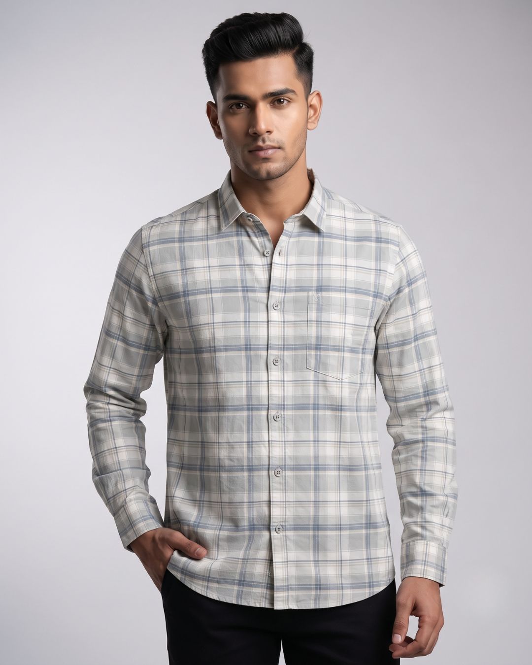 A man wearing a long-sleeve button-up shirt with a light gray and white plaid pattern, paired with black trousers, standing against a light gray studio background.