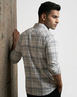 A man wearing a light beige and gray plaid long-sleeve shirt, leaning against a textured concrete pillar against a white studio background.