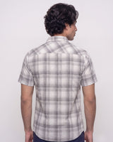 A man viewed from the back wearing a short-sleeve plaid button-up shirt in white, gray, and black, paired with dark pants, against a plain light gray studio background.