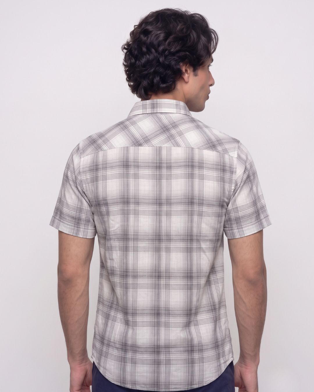 A man viewed from the back wearing a short-sleeve plaid button-up shirt in white, gray, and black, paired with dark pants, against a plain light gray studio background.