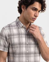 A man wearing a short-sleeve plaid button-up shirt in light gray and white, shown from the chest up against a plain light gray background.
