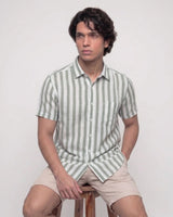 A man wearing a short-sleeve button-up shirt with vertical sage green and white stripes, paired with beige shorts, sitting on a wooden stool against a plain light background.