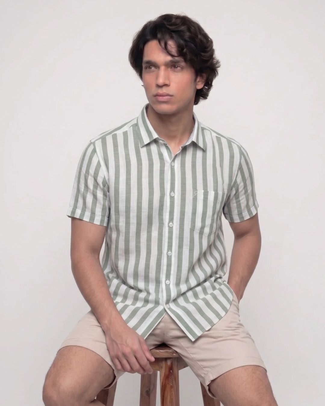 A man wearing a short-sleeve button-up shirt with vertical sage green and white stripes, paired with beige shorts, sitting on a wooden stool against a plain light background.