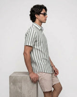 A man in profile wearing a short-sleeve button-up shirt with vertical green and white stripes, paired with beige shorts, standing next to a concrete block against a light gray background.