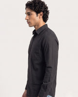 A man in profile wearing a dark charcoal gray long-sleeve button-down shirt, photographed against a clean off-white studio background.