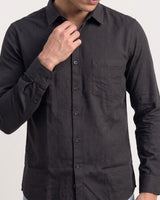 A man wearing a dark charcoal gray long-sleeve button-down shirt with a subtle geometric pattern, shown from the chest down against a light gray background.