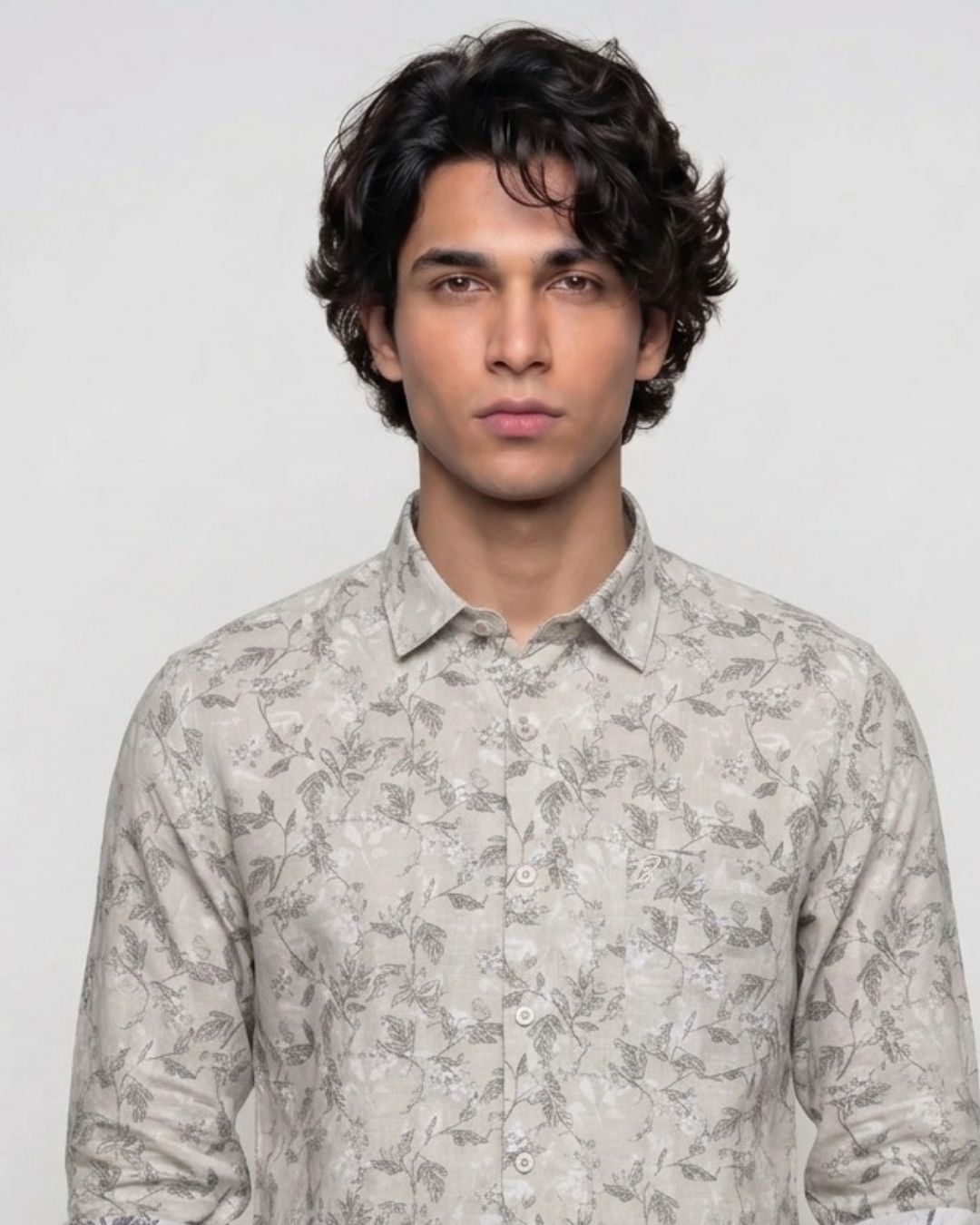 A man with dark curly hair wearing a light beige long-sleeve button-up shirt with a gray leaf pattern, photographed against a plain light gray background.