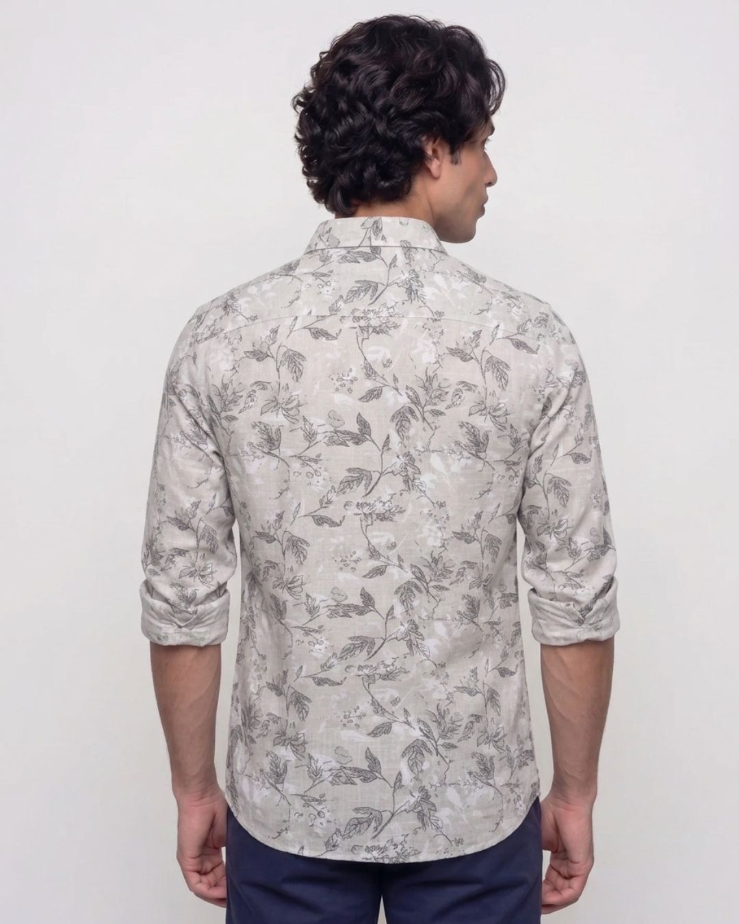 A man viewed from the back wearing a light beige long-sleeve button-up shirt with a gray floral pattern, paired with navy blue pants, against a plain white background.