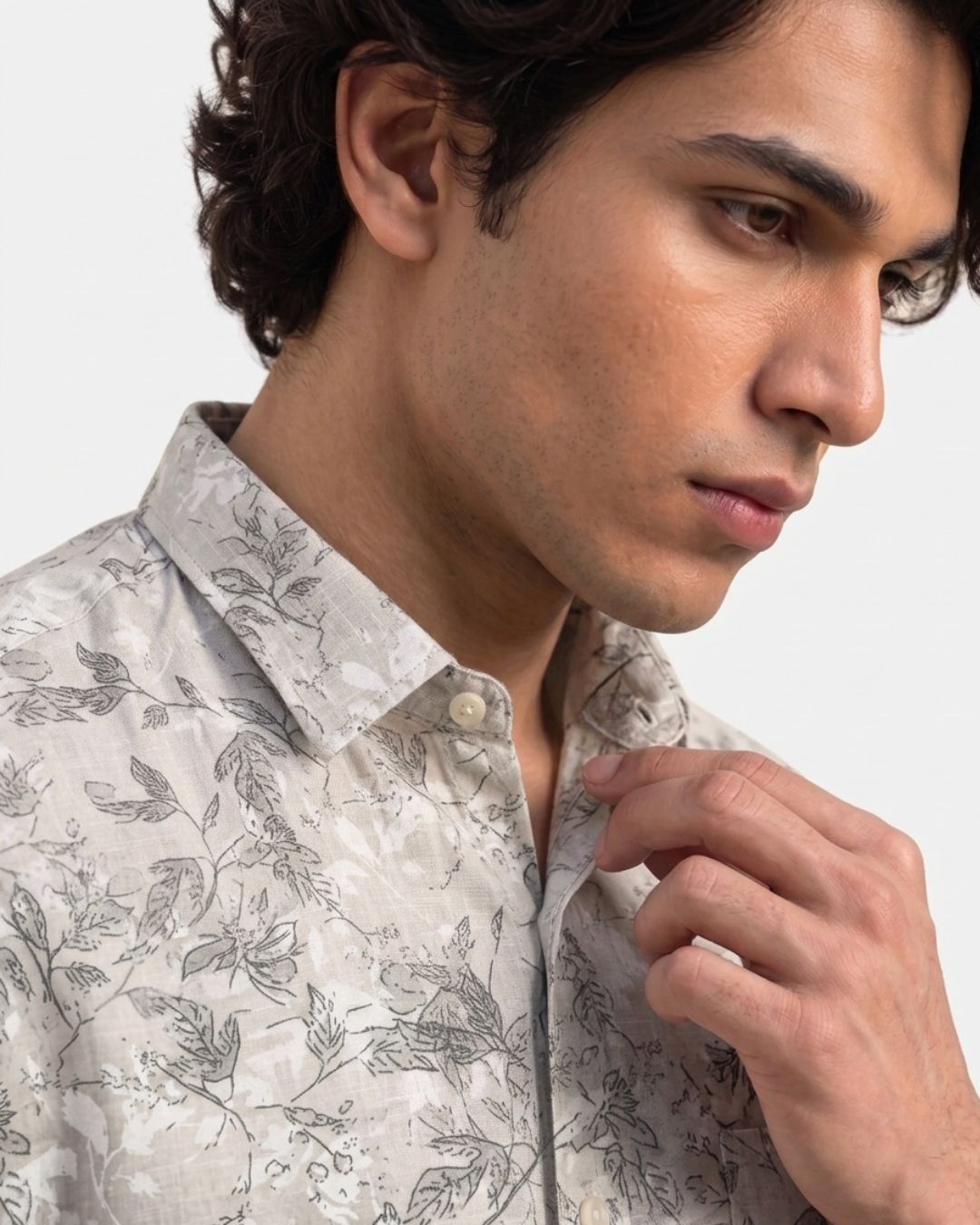 A close-up of a man wearing a light beige button-down shirt with a gray botanical leaf pattern, adjusting the collar.