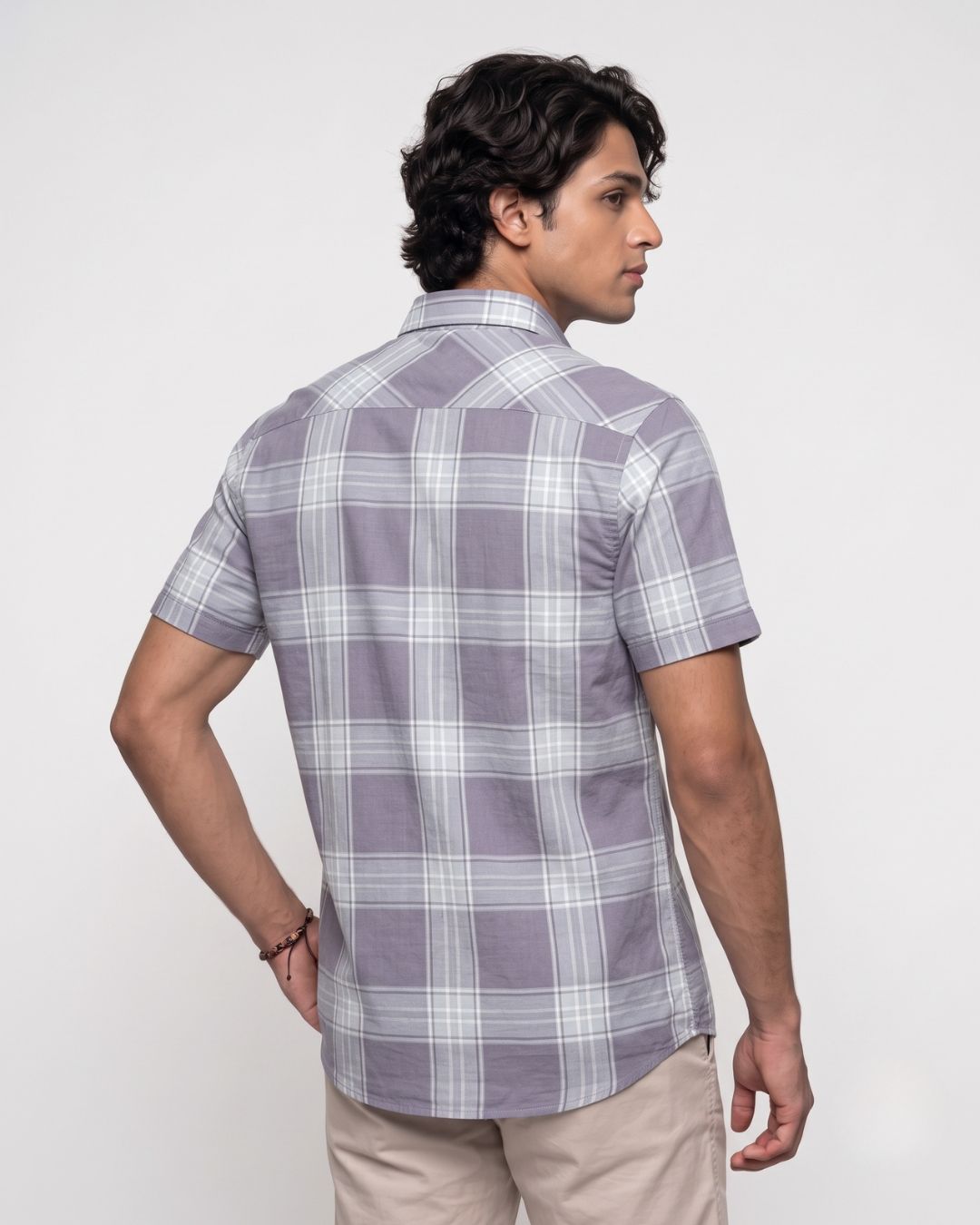 A man wearing a short-sleeve plaid button-up shirt in lavender and white, shown from a three-quarter back view against a plain light gray studio background.