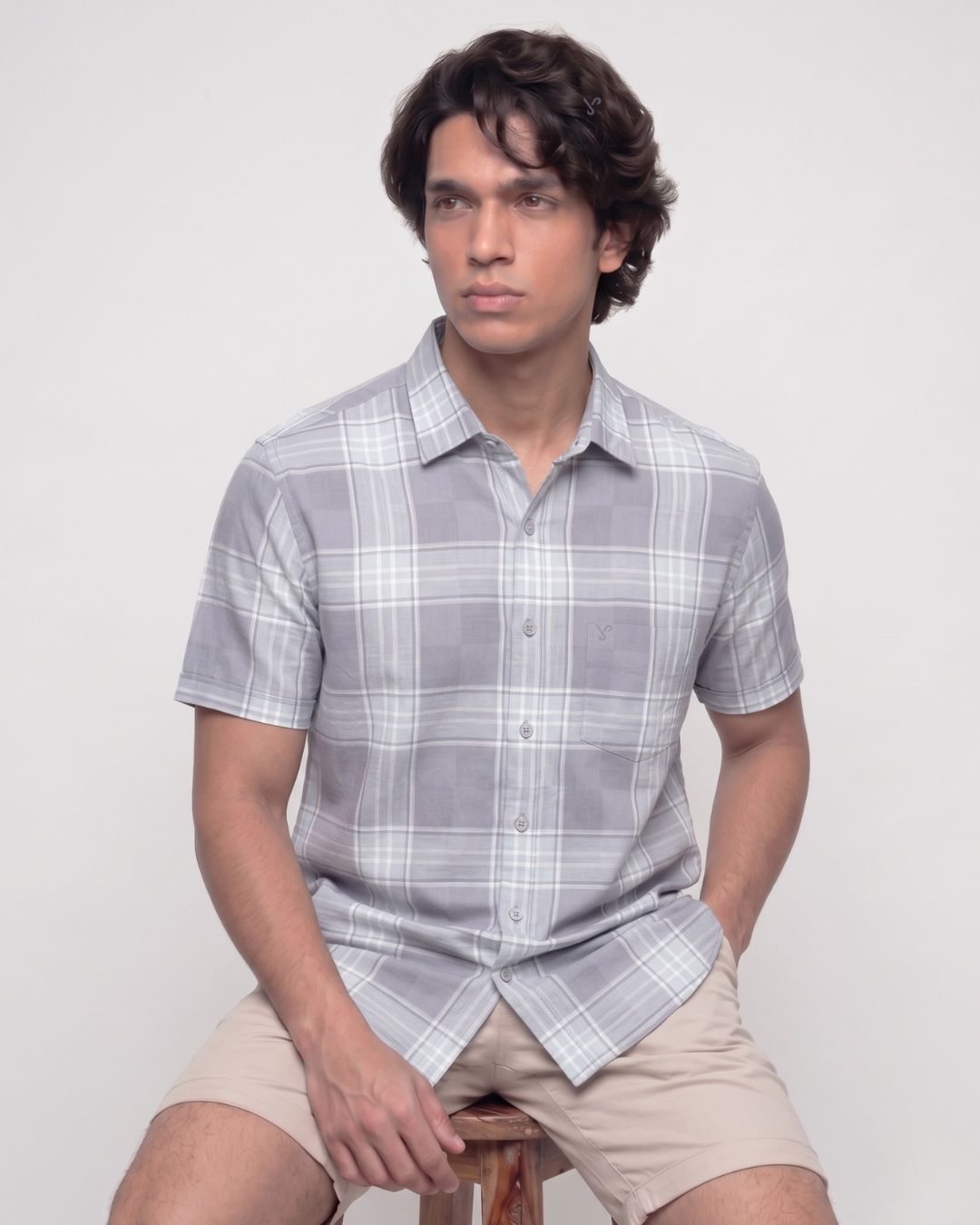 A man wearing a light gray and white plaid short-sleeve button-up shirt and beige shorts, sitting on a wooden stool against a plain light gray background.