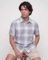 A man wearing a light gray and white plaid short-sleeve button-up shirt and beige shorts, sitting on a wooden stool against a plain light gray background.