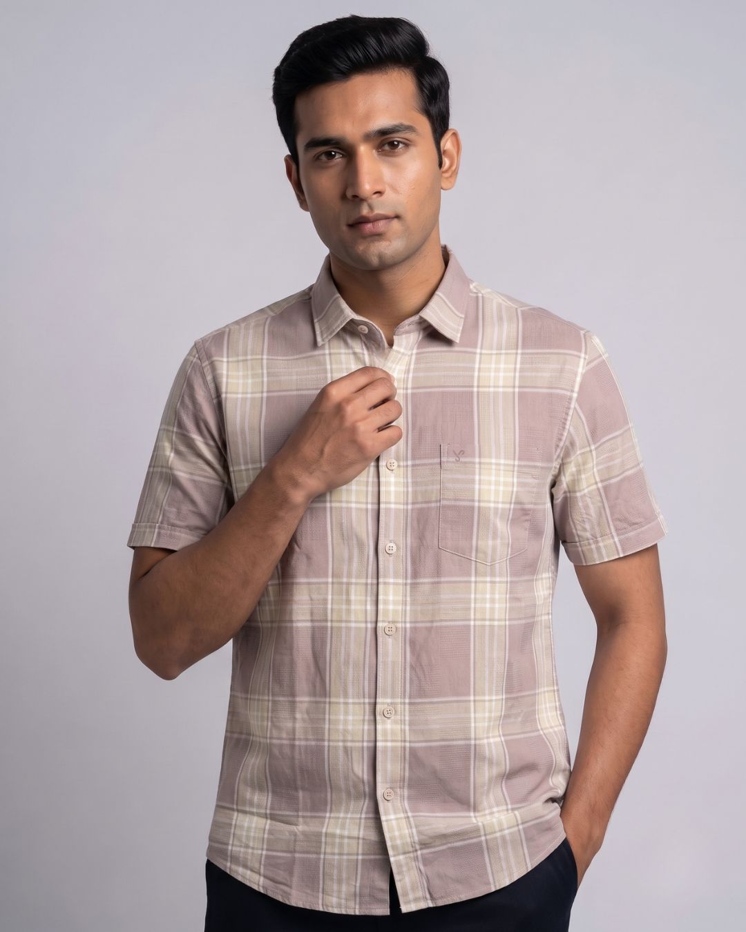 A man wearing a short-sleeve button-up plaid shirt in beige and brown tones, posed against a solid light gray studio background.