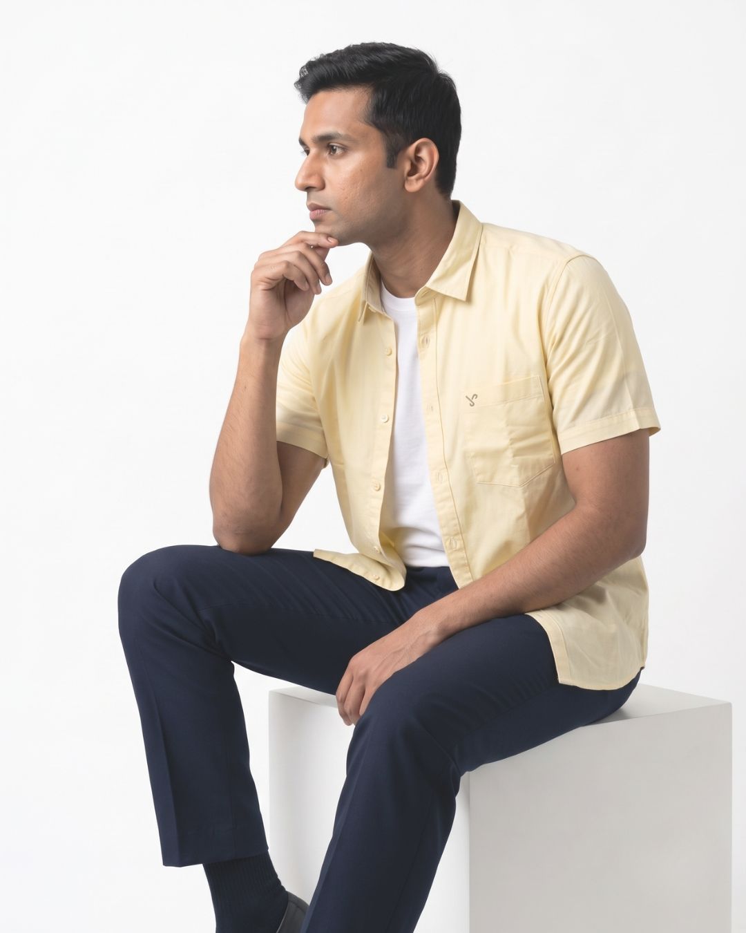 A man wearing a pale yellow short-sleeve button-up shirt over a white t-shirt and navy blue trousers, sitting on a white block against a white background.