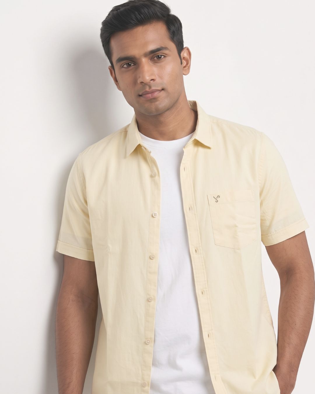 A man wearing a pale yellow short-sleeve button-up shirt over a white t-shirt, standing against a plain off-white wall.
