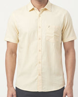 A man wearing a pale yellow short-sleeve button-up shirt with a chest pocket and a small embroidered logo, paired with dark pants against a light gray background.