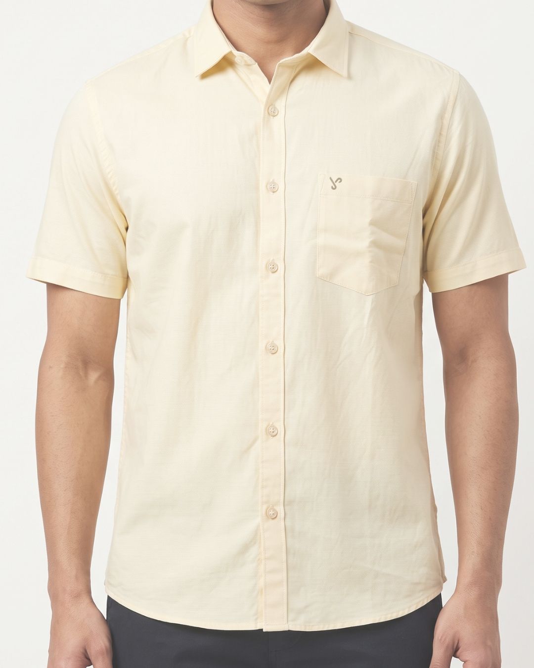A man wearing a pale yellow short-sleeve button-up shirt with a chest pocket and a small embroidered logo, paired with dark pants against a light gray background.