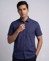 A man wearing a navy blue short-sleeve button-up shirt with thin white vertical pinstripes, standing against a light gray studio background.